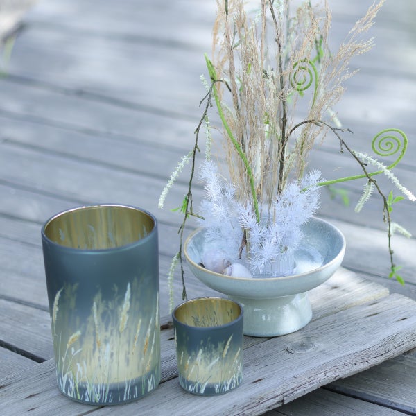 Windlichter aus Glas mit Gräser-Motiv und ein Blumengesteck in einer Keramikschale auf einer Holzoberfläche.