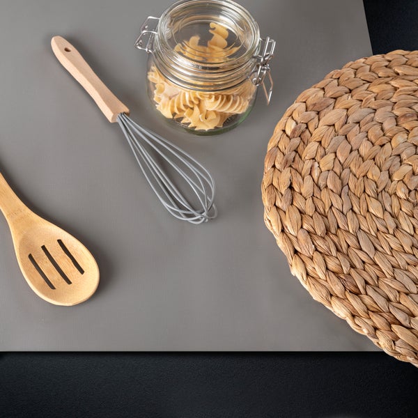 Holzlöffel, Schneebesen, Vorratsglas mit Nudeln und geflochtenes Platzset auf einer grauen Antirutschmatte.