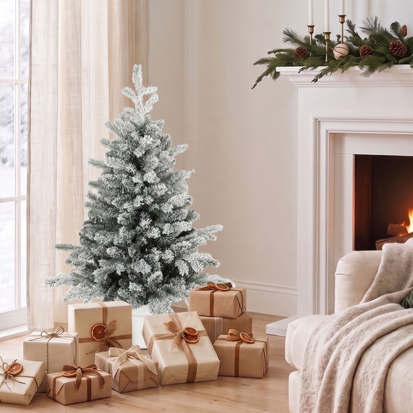 Ein künstlicher, verschneiter Weihnachtsbaum steht neben einem Kamin in einem gemütlichen Wohnzimmer, umgeben von weihnachtlich verpackten Geschenken.