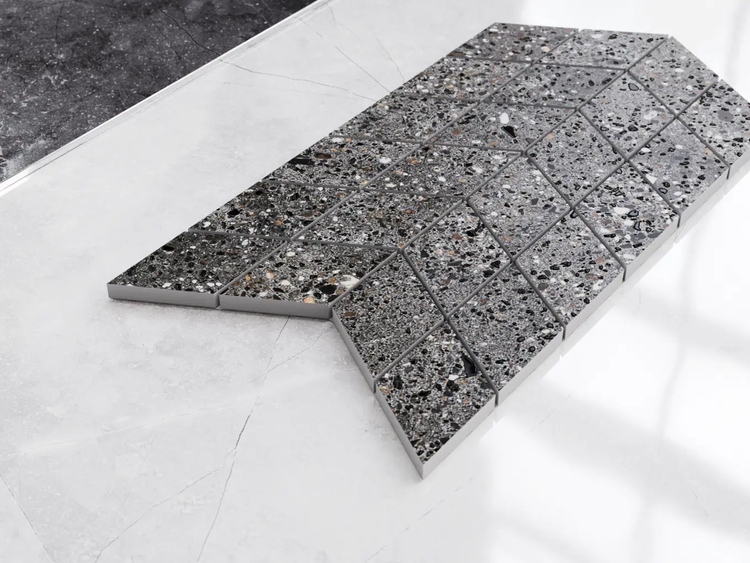 Mosaikfliese in Terrazzo-Optik, Chevron-Muster aus rautenförmigen Steinchen, grau gesprenkelt.