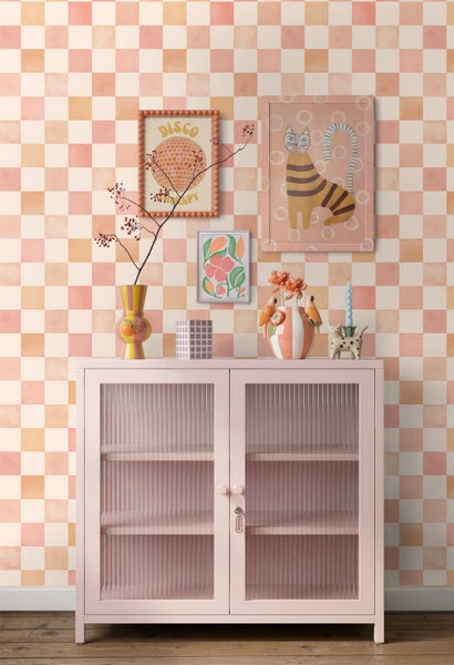 Vliestapete mit pastellfarbenem Schachbrettmuster hinter einer hellrosa Anrichte mit Riffelglas und bunten Kunstdrucken an der Wand.