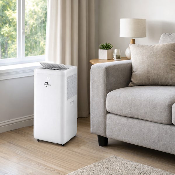 Mobiles Klimagerät mit Sannover Logo und Digitalanzeige in einem hellen, modern eingerichteten Wohnzimmer neben einem grauen Sofa und Fenster.