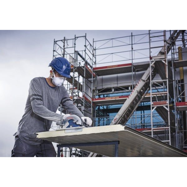 Ein Mann mit Schutzhelm, Schutzbrille und Atemschutzmaske sägt mit einer Handkreissäge eine Platte auf einem Tisch. Im Hintergrund befindet sich ein Baugerüst.