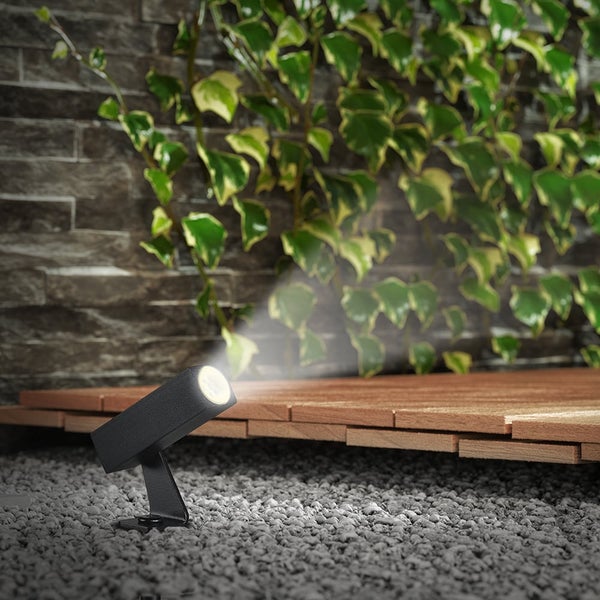 Moderner schwarzer LED-Außenstrahler auf Kies beleuchtet eine Szene mit Holzterrasse und einer Steinwand mit Efeu.