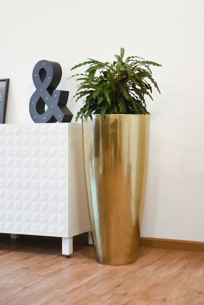 Hoher Pflanzkübel in Goldoptik mit grüner Pflanze in einem hellen, modernen Wohnraum neben einem weißen Sideboard.