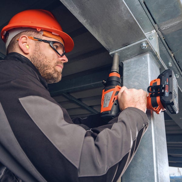 Ein Arbeiter mit Schutzhelm, Schutzbrille und Gehörschutz verwendet einen Akku-Schlagschrauber an einer Metallkonstruktion.