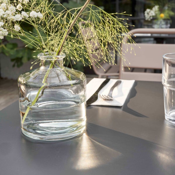 Kleine runde Glasvase mit Wasser und zarten weißen Blumen auf einem grauen Tisch im Freien mit Besteck und einem Trinkglas.