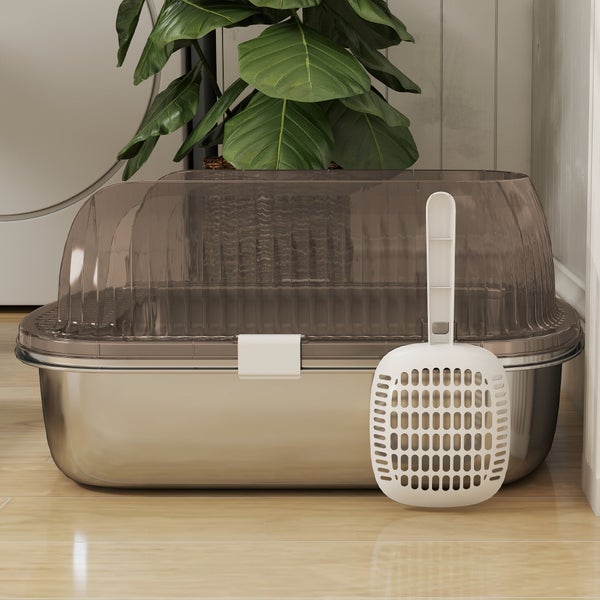Katzentoilette mit Edelstahl-Wanne, transparenter Haube und Streuschaufel auf hellem Holzboden neben einer Zimmerpflanze.