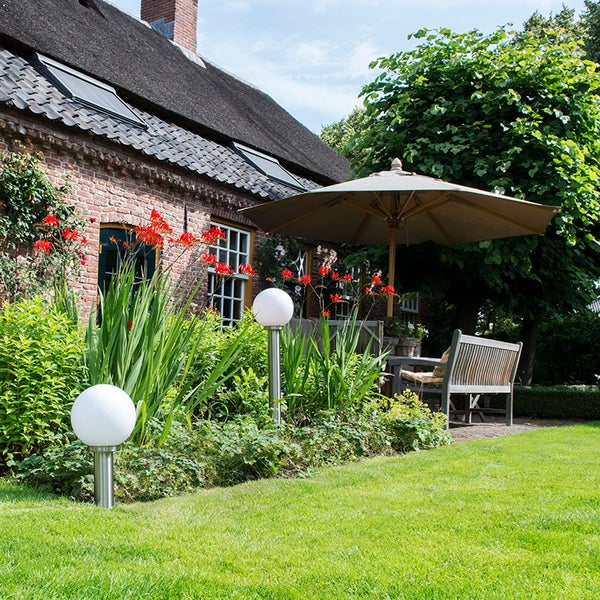 Zwei kugelförmige Gartenleuchten in einem Blumenbeet neben einer Rasenfläche vor einem Haus mit Sonnenschirm und Gartenbank.