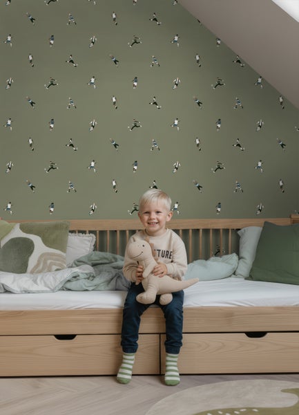 Tapete mit Fußballspieler-Motiv im hellen Kinderzimmer, Kind sitzt auf Holzbett mit Dinosaurier-Kuscheltier und Kissen.