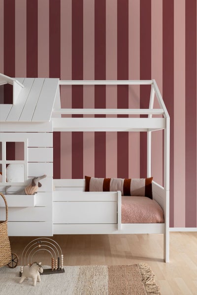 Kinderzimmer mit weißem Hausbett aus Holz vor einer vertikal gestreiften Tapete in Rottönen, Teppich und Holzspielzeug auf hellem Boden.