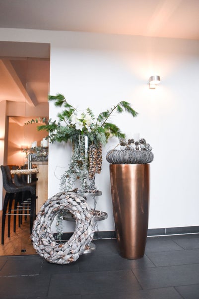 Festliche Innendekoration mit großer bronzefarbener Bodenvase, Etagere mit Tannengrün und Blumen sowie geflochtenen Kränzen in einem hellen Raum.