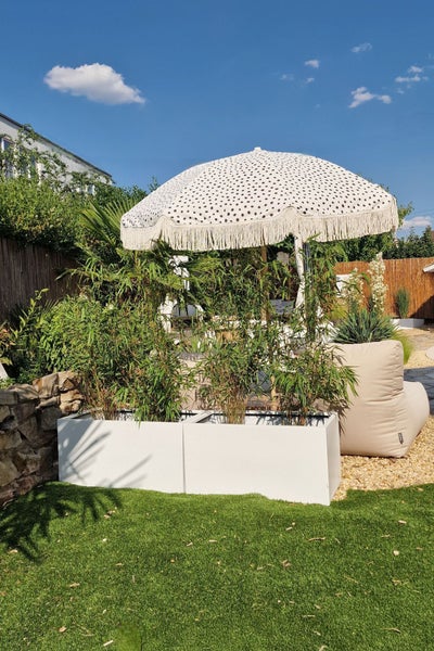Moderne Gartenszene mit zwei rechteckigen weißen Pflanzkübeln mit Bambus, einem gemusterten Sonnenschirm und einem beigen Sitzsack.