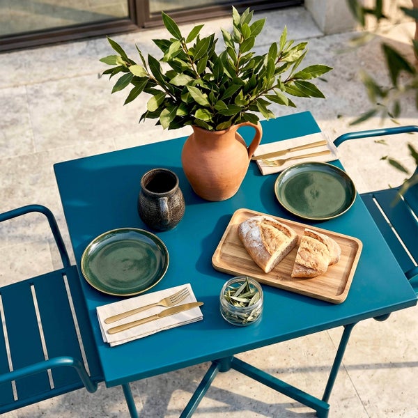 Gedeckter Gartentisch aus Metall für zwei Personen mit Tellern, Besteck, Brot auf einem Holzbrett und einem Terrakotta-Krug auf einer Terrasse.
