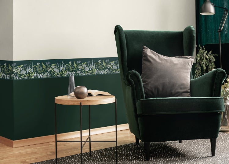 Florale Tapetenborte an einer zweifarbigen Wand in einem Wohnzimmer mit grünem Samtsessel und rundem Beistelltisch aus Holz.