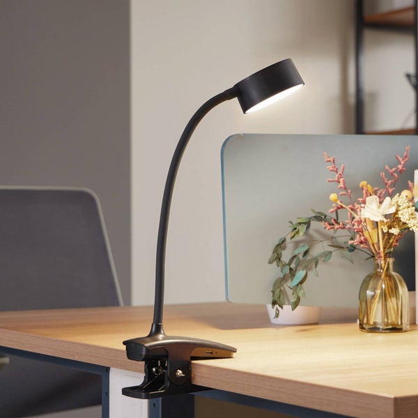 Schwarze LED-Klemmleuchte mit flexiblem Hals und USB-Anschluss, befestigt an einem hellen Schreibtisch aus Holz in einem modernen Büro.