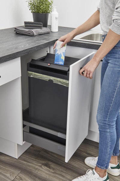 Organisiertes, ausziehbares Abfallsystem zur Mülltrennung im weißen Küchenunterschrank mit einer Person, die eine Verpackung entsorgt.