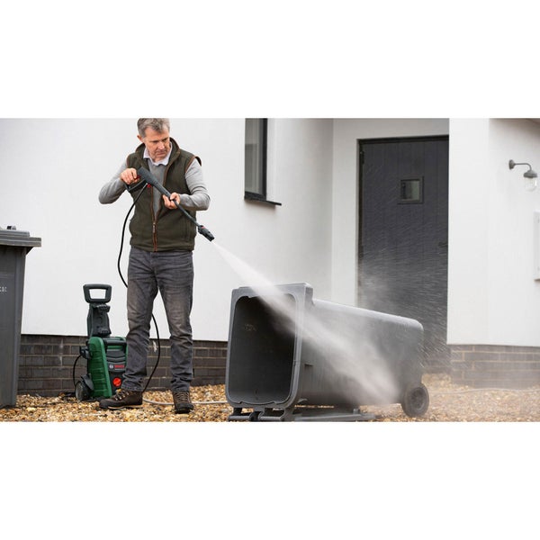 Ein Mann reinigt eine liegende Mülltonne im Außenbereich mit einem Bosch Hochdruckreiniger auf einer Kiesfläche vor einem Haus.