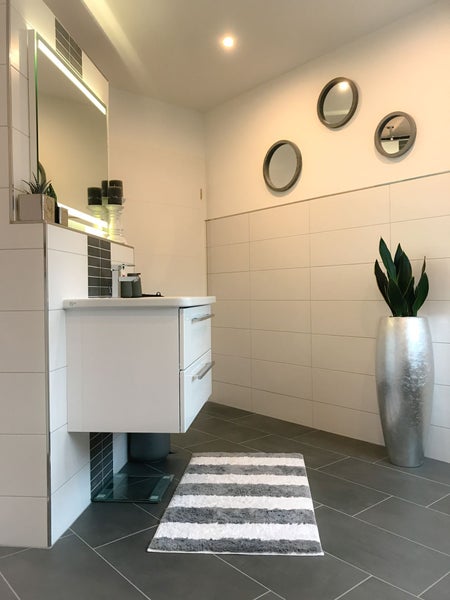 Modernes Badezimmer mit weißem Waschtischunterschrank, Spiegel mit Beleuchtung, silberner Bodenvase mit Pflanze und gestreiftem Badeteppich.