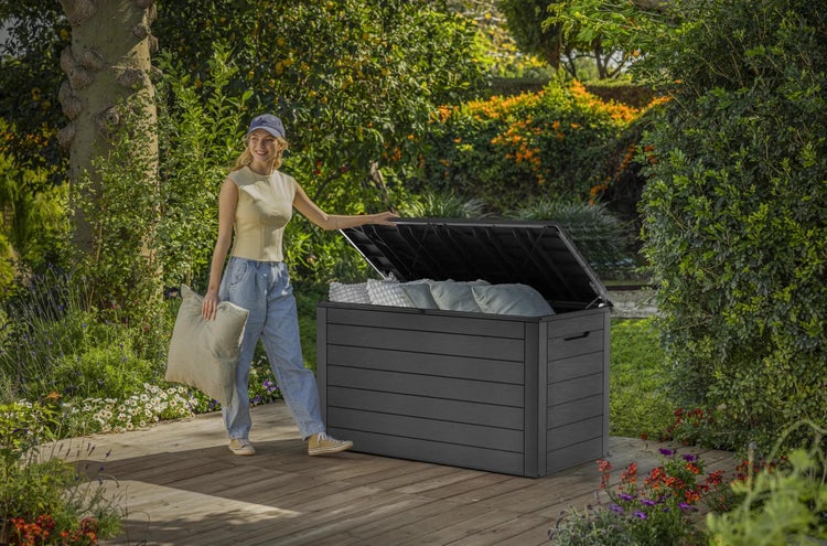 Große dunkelgraue Garten-Auflagenbox in Holzoptik auf einer Holzterrasse im üppigen Garten, eine Person legt helle Kissen hinein.