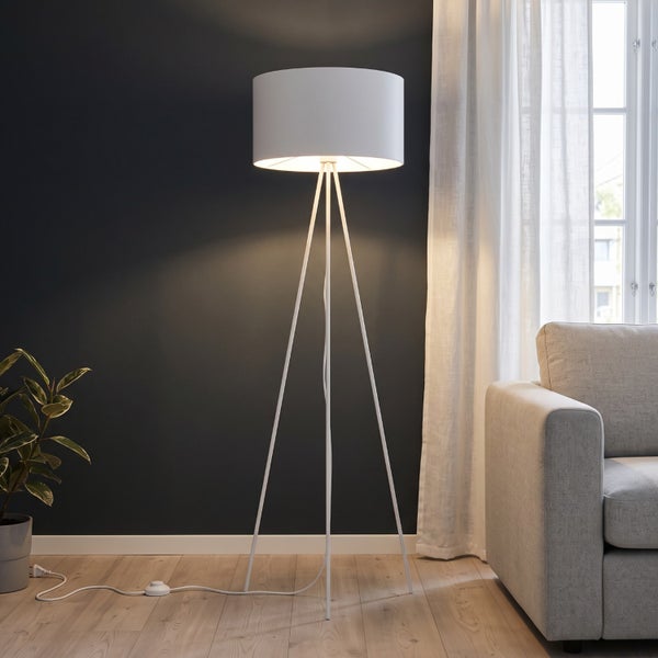 Moderne Stehlampe mit zylindrischem Lampenschirm auf einem dreibeinigen Stativgestell in einem Wohnzimmer mit Sofa und Vorhängen.