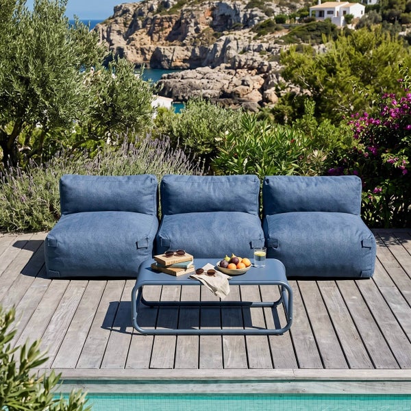 Blaue Lounge-Sitzsäcke und niedriger Metalltisch auf einer Holzterrasse mit Blick auf das Meer, umgeben von mediterranen Pflanzen.