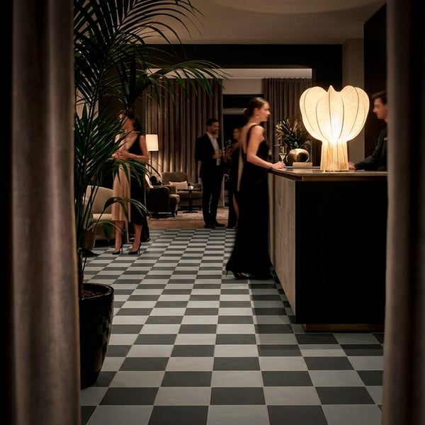 Elegantes Interieur mit Fliesenboden im Schachbrettmuster, einer Zimmerpflanze und einer dekorativen Tischleuchte in einer Hotelbar.