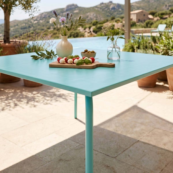 Quadratischer Metall-Gartentisch auf einer sonnigen Terrasse am Pool, dekoriert mit Servierbrett, Oliven und Blumenvasen.