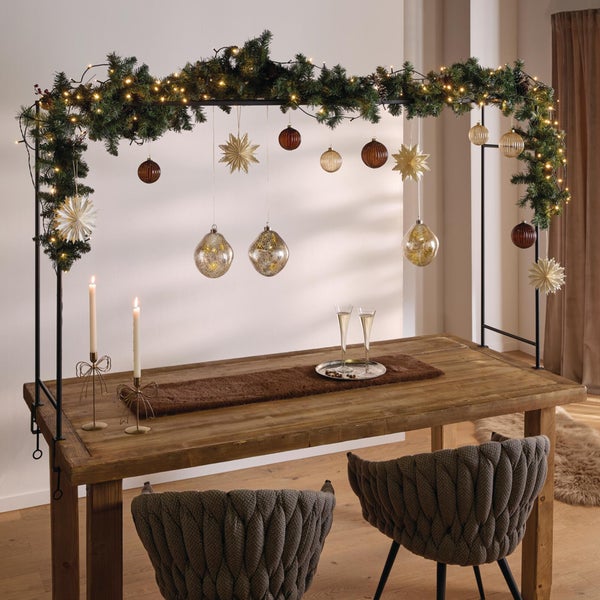 Ein weihnachtlich dekorierter Tischaufsatz aus Metall über einem rustikalen Holztisch mit Lichterkette, Christbaumkugeln und Sternenanhängern.