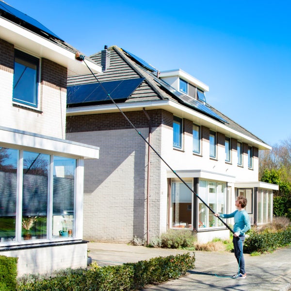 Eine Person reinigt die Dachrinne eines zweistöckigen Hauses mit einem langen, ausziehbaren Teleskopstiel und einer Reinigungsbürste.