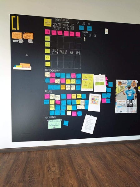 Große schwarze Wandtafel zur Organisation im Büro mit bunten Notizzetteln, Zeitplänen und Postern über einem Holzboden.