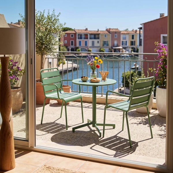 Balkonset mit zwei Metallstühlen und einem runden Metalltisch auf einem hellen Balkon mit Pflanzen und Blick auf bunte Häuser am Wasser.
