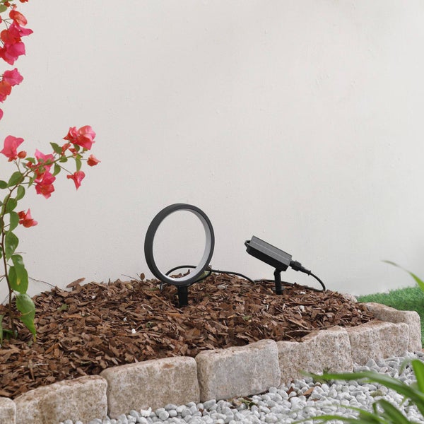 Ringförmige LED-Außenleuchte und separates Solarpanel in einem mit Mulch gefüllten Gartenbeet mit Steinumrandung.