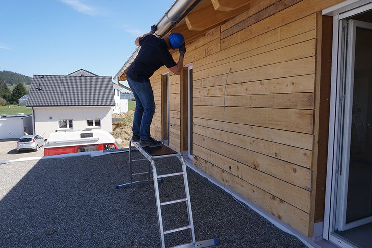 Person auf einer Leiter bei Arbeiten an einer Holzfassade mit Dachrinne