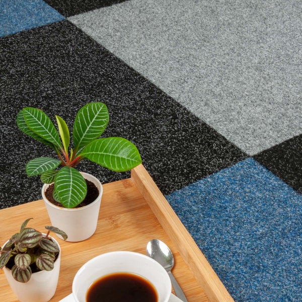 Quadratische Teppichfliesen in Dunkelgrau, Hellgrau und Blau mit einem Holztablett, zwei Zimmerpflanzen und einer Tasse Kaffee.