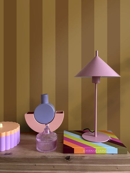 Moderne Innenraumszene mit einer rosa Tischleuchte auf einem bunten Buch, dekorativen Vasen und einem Glasgefäß auf Holz vor einer gestreiften Wand.