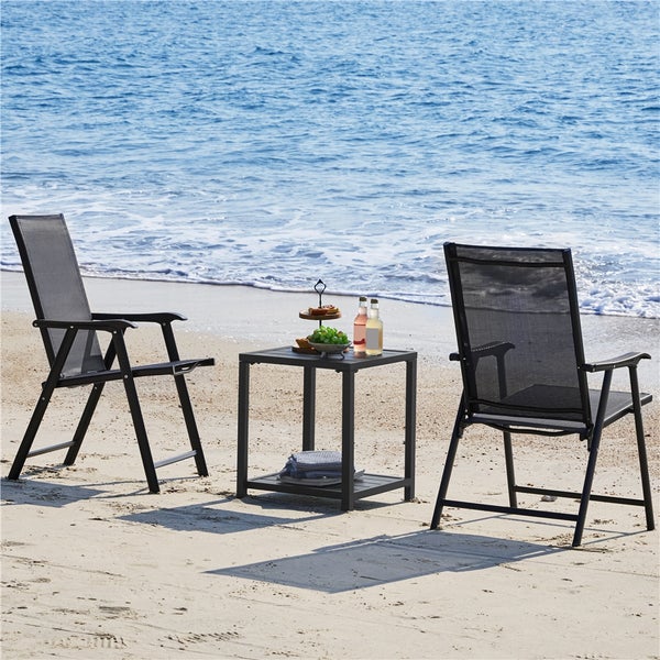 Zwei dunkelgraue Gartenklappsessel aus Metall und ein quadratischer Beistelltisch auf einem sonnigen Sandstrand am Meer.