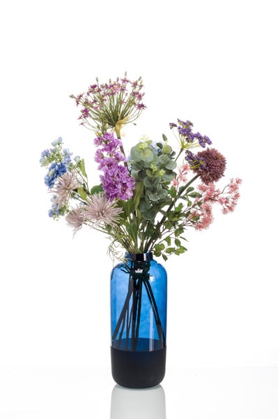 Künstlicher Blumenstrauß mit lila, blauen und rosa Kunstblumen sowie grünen Zweigen in einer blauen Glasvase.