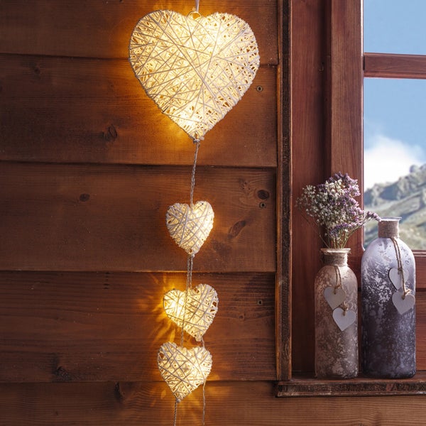 Dekoratives Arrangement mit einer Herzchen-Lichterkette, die an einer Holzwand hängt, neben einem Fenster mit Vasen auf dem Sims