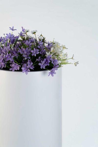 Künstliche lila und weiße Blumen in einem weißen zylindrischen Pflanzgefäß.