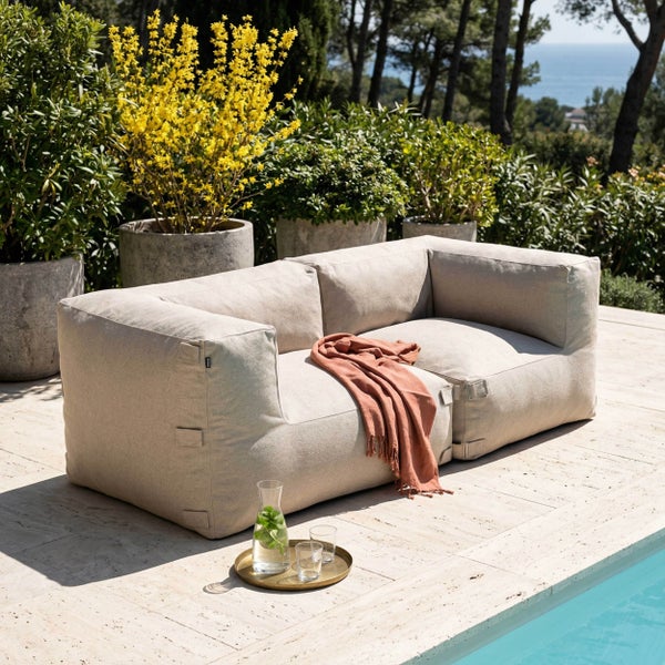 Modernes Gartensofa aus beigem Stoff auf einer Terrasse am Pool, mit dekorativer Decke und Tablett, umgeben von Pflanzen.
