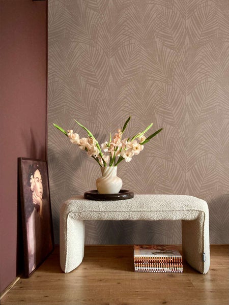 Wohnraum mit Vliestapete mit Palmenblattmuster, davor eine Sitzbank aus Bouclestoff, eine Vase mit Gladiolen und ein gerahmtes Bild auf dem Boden.