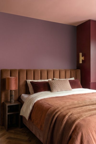 Modernes Schlafzimmer mit gepolstertem Kopfteil, farbig gestalteten Wänden, Nachttischlampe und dekorativen Textilien auf Parkettboden.