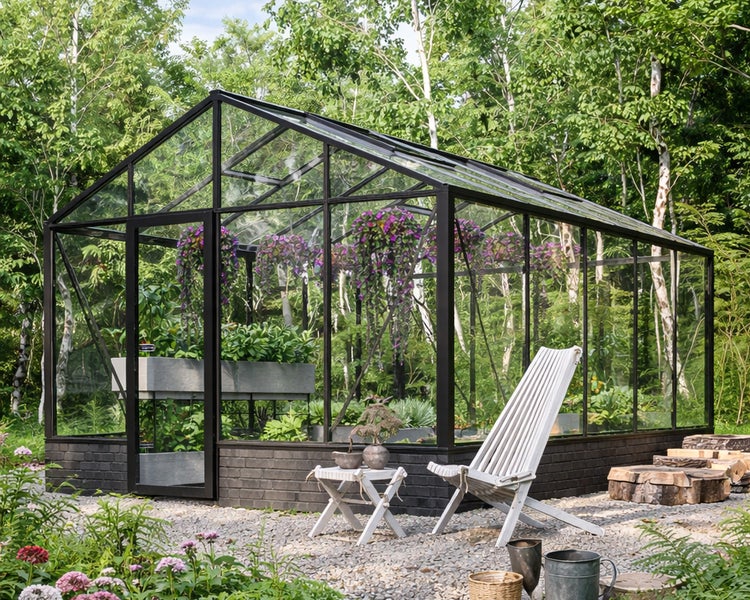 Modernes Gewächshaus mit schwarzem Rahmen auf Steinsockel. Innen lila Blumen und grüne Pflanzen. Außen Kiesplatz mit weißem Stuhl vor Bäumen.