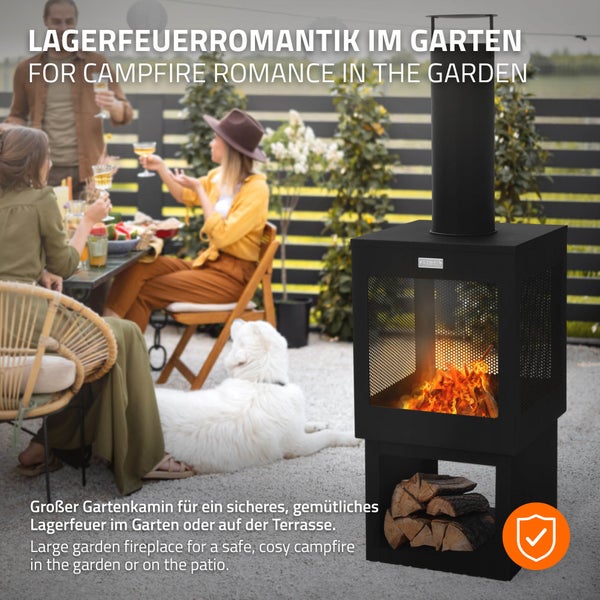 Moderner schwarzer Gartenkamin mit Schornstein und Holzfach auf einer Terrasse, im Hintergrund eine Gruppe von Personen an einem Tisch im Garten.