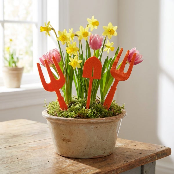 Frühlingsdekoration mit Narzissen und Tulpen im Tontopf dekoriert mit kleinen Gartenwerkzeugen aus Filz auf einem Holztisch.
