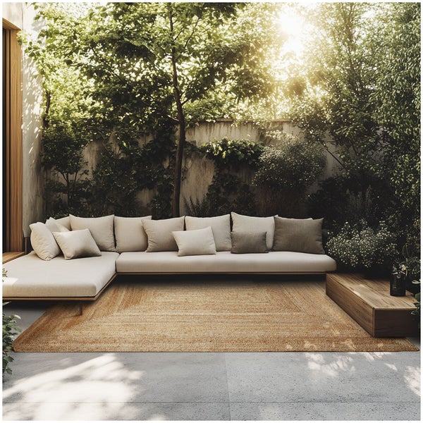 Moderne Loungegarnitur mit Ecksofa und Kissen auf einer Terrasse vor einer Mauer mit Gartenpflanzen und einem gewebten Teppich.