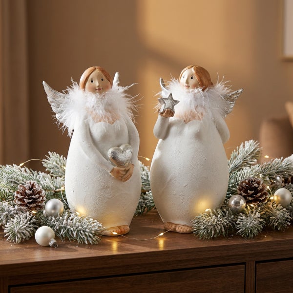 Zwei dekorative Weihnachtsengel Figuren mit Federkragen, einer mit Herz und einer mit Stern, umgeben von Tannenzweigen und Lichterkette.