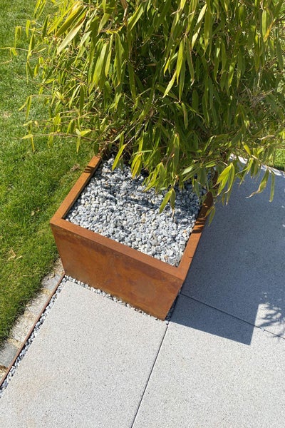 Quadratischer Pflanzkübel aus Cortenstahl mit Rostoptik, bepflanzt mit Bambus und Kiesschüttung auf einer Terrasse.
