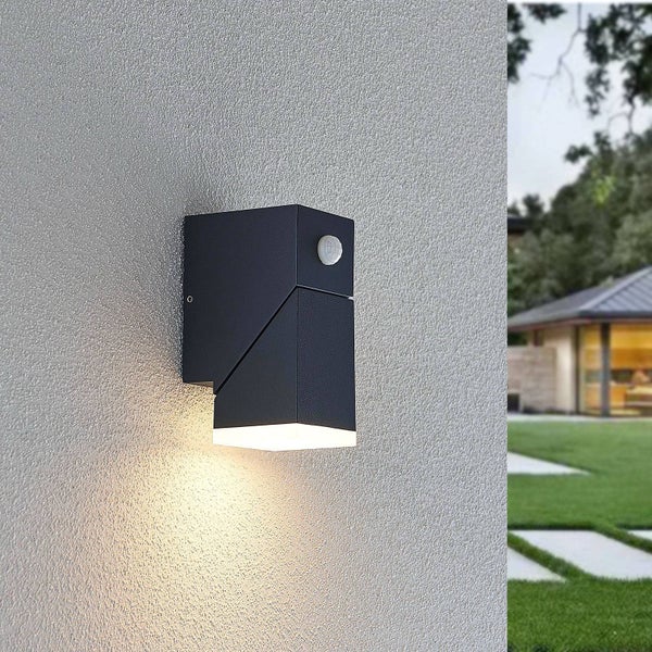 Beleuchtete moderne anthrazitfarbene Außenwandleuchte mit Bewegungsmelder an einer Hauswand, im Hintergrund ein Garten mit Rasen und ein Gebäude.